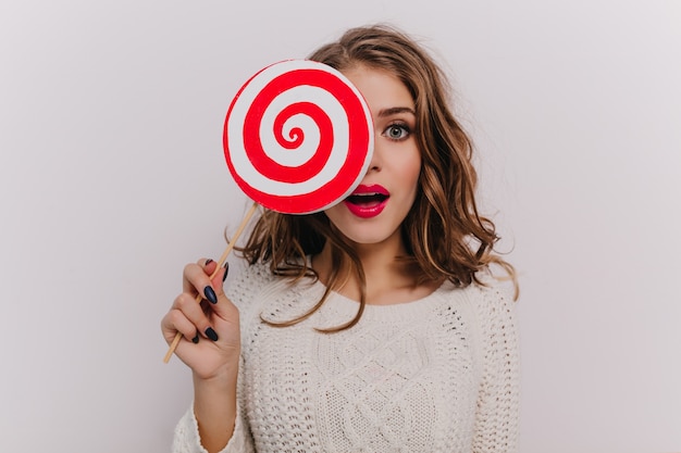 Retrato de close-up de uma garota cacheada de olhos cinza com lábios vermelhos cobrindo o rosto com um doce enorme