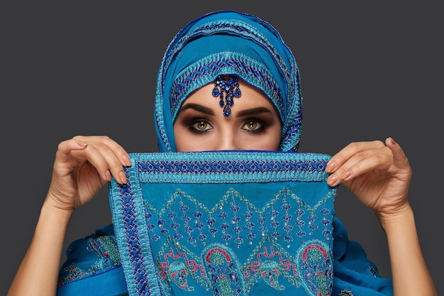 Retrato de close-up de uma jovem fêmea com olhos esfumaçados, vestindo um elegante hijab azul decorado com lantejoulas e joias. ela está cobrindo o rosto com um xale e olhando para a câmera em um fundo escuro