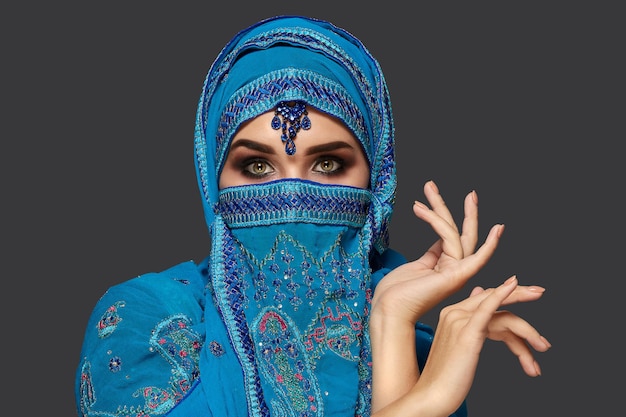 Retrato de close-up de uma menina bonita com encantadores olhos esfumaçados, vestindo um hijab azul decorado com lantejoulas e joias. Ela está gesticulando e olhando para a câmera em um fundo escuro. Emoções humanas