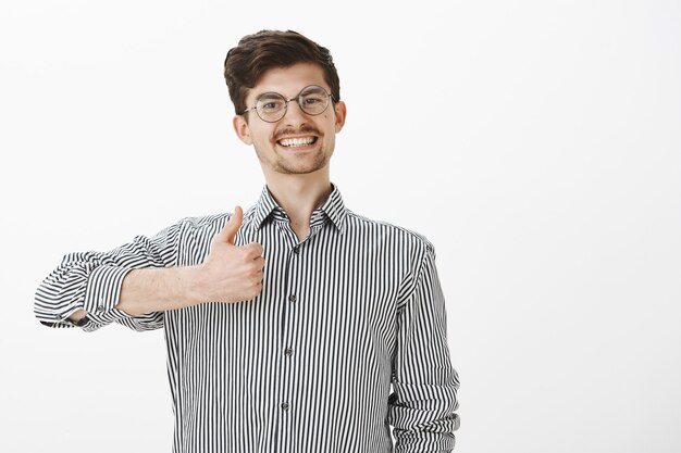 Retrato de colega de trabalho amigável ativo e positivo de óculos redondos, sorrindo alegremente enquanto mostra os polegares para cima, estando pronto para qualquer tipo de trabalho, aprovando e dizendo que gosta de ideias sobre parede cinza