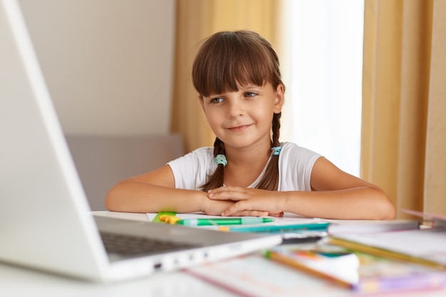 Retrato de colegial com cabelo escuro e rabo de cavalo, sentado à mesa na sala de estar. professora ouvinte online, tendo aula a distância em quarentena, usufruindo de educação à distância.