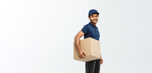 Foto grátis retrato de conceito de entrega de entregador afro-americano feliz em pano azul andando para enviar um pacote de caixa ao cliente isolado no espaço de cópia de fundo do estúdio cinza