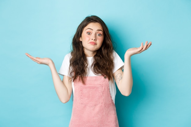 Retrato de confusa linda menina morena encolhendo os ombros, não sei de nada, não posso dizer, indeciso contra um fundo azul.