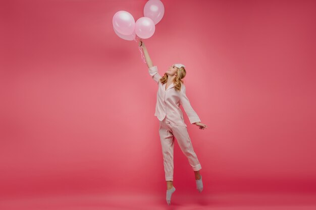 Retrato de corpo inteiro da adorável garota despreocupada, na ponta dos pés com balões. Foto interna da senhora encaracolada em pijamas rosa e máscara, saltando com um sorriso.