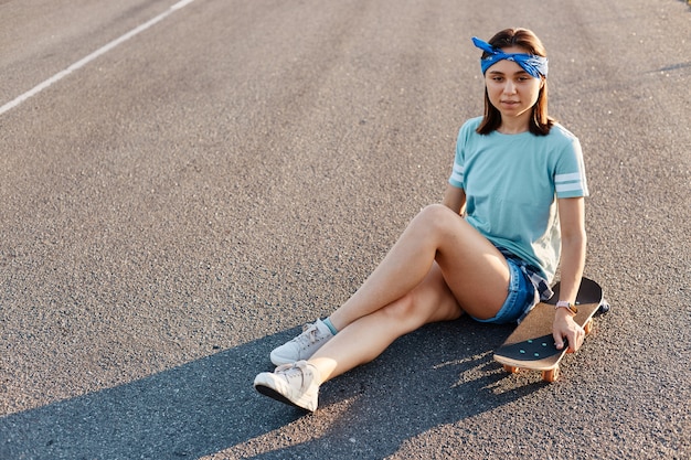 Retrato de corpo inteiro da bela mulher vestindo camiseta casual, curto e faixa de cabelo, sentado na estrada de asfalto com skate, olhando para longe com expressão facial sonhadora.