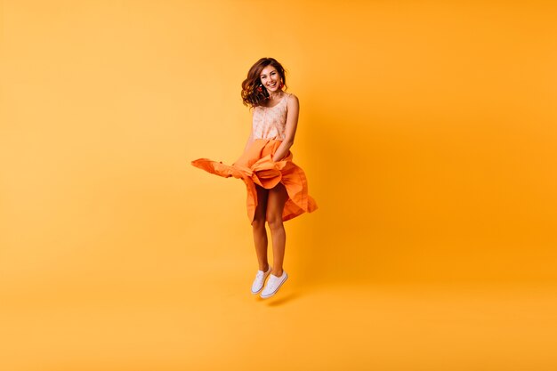 Retrato de corpo inteiro de elegante senhora elegante pulando no estúdio. Linda garota ruiva com saia laranja se divertindo.