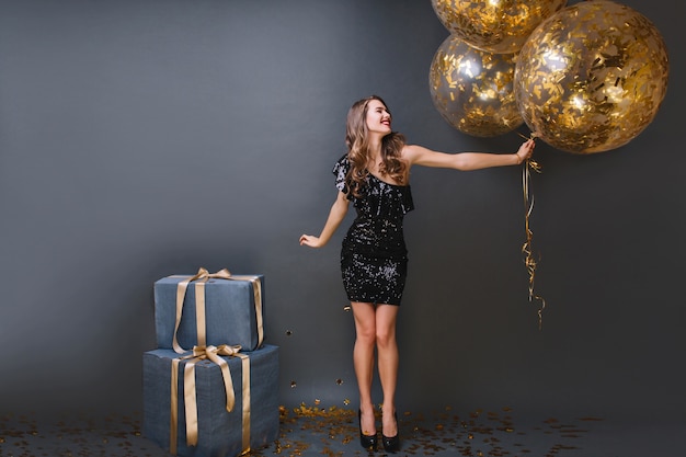 Retrato de corpo inteiro de garota europeia refinada usa vestido preto na festa de aniversário. feliz senhora de cabelos compridos com balões mal pode esperar para abrir os presentes.