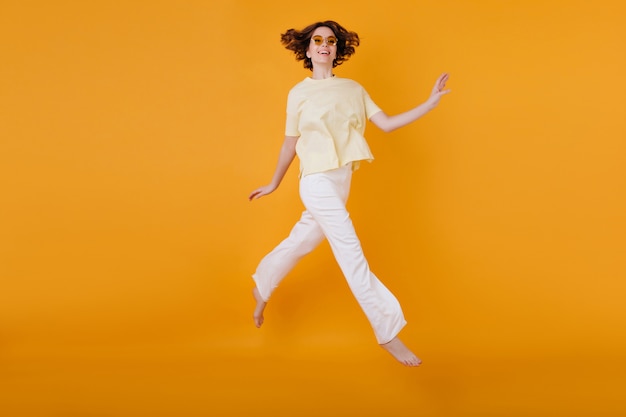 Retrato de corpo inteiro de garota feliz em camiseta amarela e calça branca correndo na parede laranja. mulher caucasiana maravilhosa dançando com prazer.