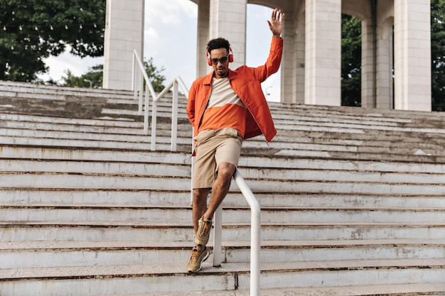 Retrato de corpo inteiro de homem moreno encaracolado de pele escura em shorts bege e jaqueta laranja posando e se divertindo nas escadas do lado de fora jovem de óculos de sol ouve música em fones de ouvido