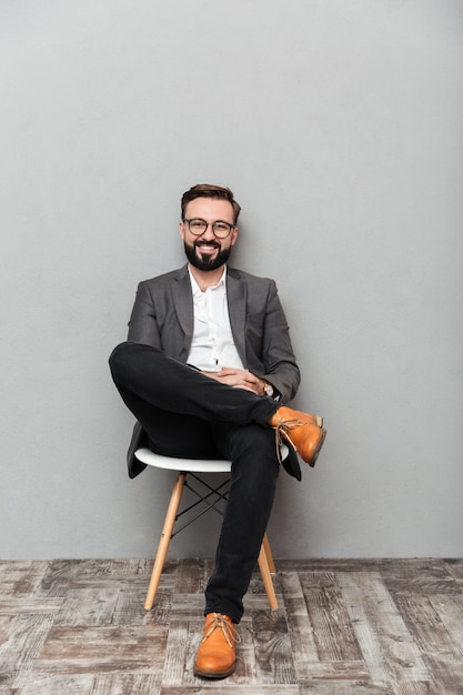 Retrato de corpo inteiro de homem relaxado em casual sentado na cadeira no escritório e sorrindo para a câmera, isolada sobre cinza