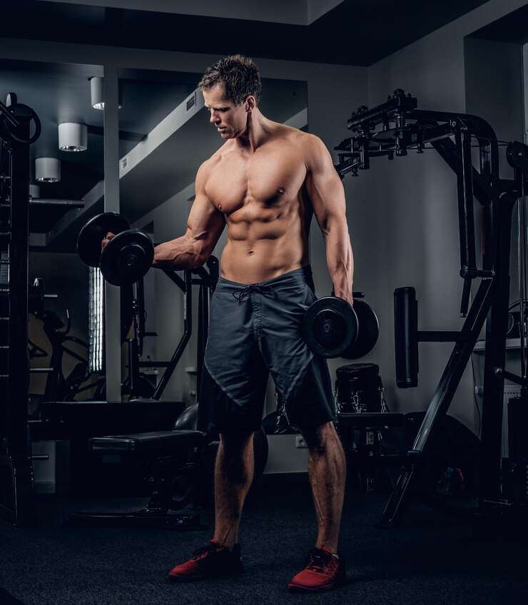 Foto grátis retrato de corpo inteiro de homem sem camisa atlético fazendo exercícios de bíceps com halteres em um clube de ginástica.