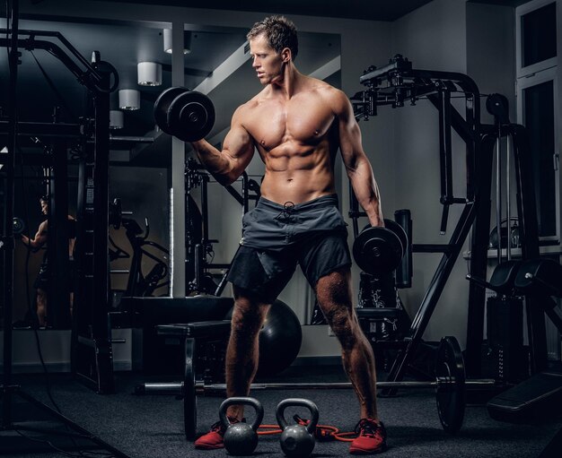 Retrato de corpo inteiro de homem sem camisa atlético fazendo exercícios de bíceps com halteres em um clube de ginástica.
