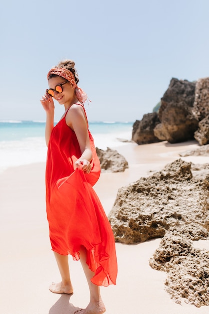Foto grátis retrato de corpo inteiro de jovem alegre, brincando, olhando por cima do ombro na praia. menina bonita em um vestido longo vermelho, andando na praia.