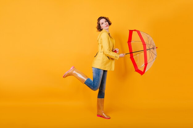 Retrato de corpo inteiro de menina bem torneada em sapatos de borracha dançando com guarda-sol vermelho. Senhora encaracolada na jaqueta amarela em pé sobre uma perna e segurando o guarda-chuva.