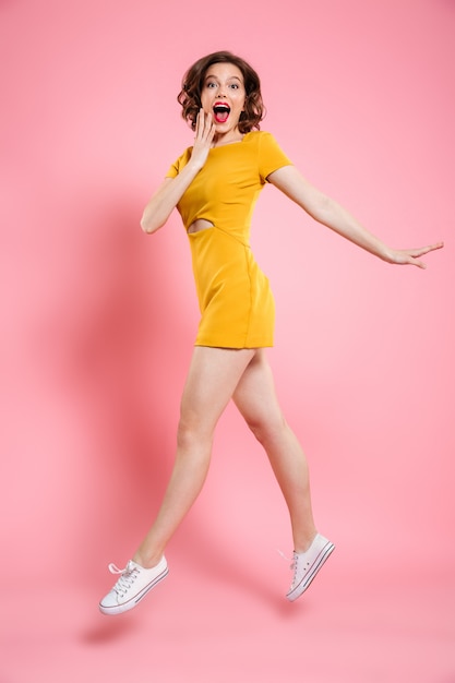 Retrato de corpo inteiro de mulher feliz saiu no elegante vestido amarelo ao saltar sobre rosa