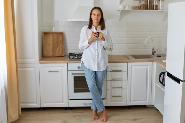 Retrato de corpo inteiro de mulher tomando café pela manhã em casa, em pé perto do conjunto de cozinha, desfrutando de uma bebida quente em casa, vestindo jeans e camisa branca.