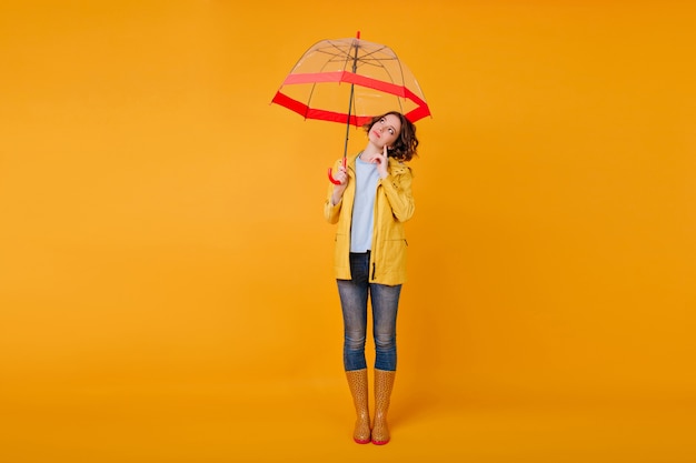 Retrato de corpo inteiro de pensativa menina romântica em pé na parede amarela sob o guarda-sol vermelho. foto de estúdio do elegante modelo feminino em jeans e sapatos de outono, olhando para longe enquanto posava com guarda-chuva.