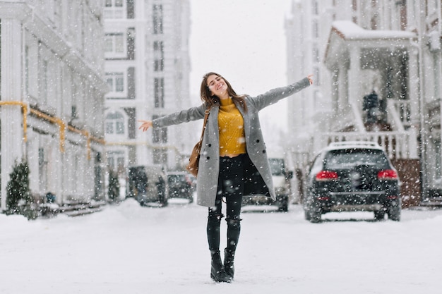 Retrato de corpo inteiro de romântica senhora europeia usa casaco longo em dia de neve. Foto ao ar livre de inspirada mulher morena, aproveitando o tempo livre na cidade de inverno.