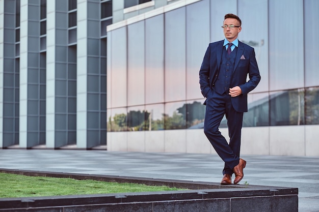 Retrato de corpo inteiro de um homem confiante em um terno elegante posando com a mão no bolso contra um fundo de arranha-céu.