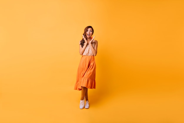 Retrato de corpo inteiro de uma garota incrível com roupa de verão brilhante. mulher morena alegre posando em amarelo com um sorriso de surpresa.