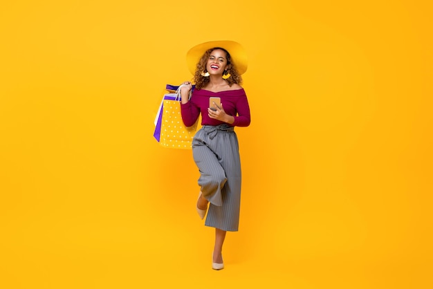 Retrato de corpo inteiro de uma jovem e atraente afro-americana feliz segurando sacolas de compras e celular com uma perna levantada em fundo amarelo estúdio isolado