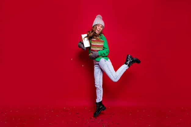 Retrato de corpo inteiro de uma jovem elegante e feliz vestida de suéter verde, calça jeans, botas pretas e boné de inverno cinza pulando com presentes de ano novo em fundo vermelho isolado