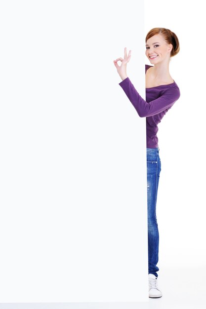 Retrato de corpo inteiro de uma jovem olhando para fora por causa de um outdoor de publicidade com um gesto de ok