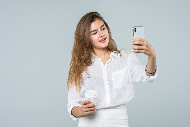 Retrato de corpo inteiro de uma menina sorridente feliz usando telefone celular em pé e segurando a xícara de café sobre fundo branco