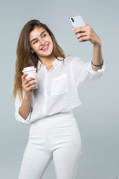 Retrato de corpo inteiro de uma menina sorridente feliz usando telefone celular em pé e segurando a xícara de café sobre fundo branco