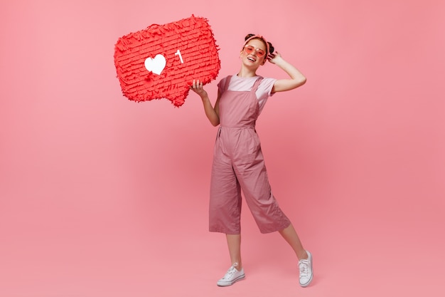 Retrato de corpo inteiro de uma mulher ativa em roupa rosa e óculos de sol, segurando como cartaz e pulando no fundo isolado.