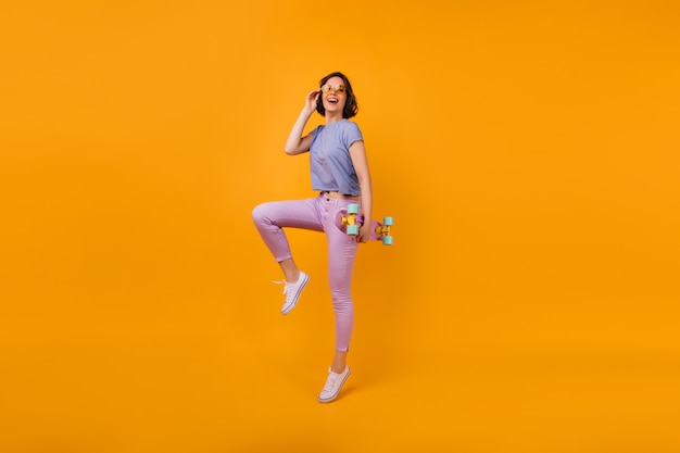 Retrato de corpo inteiro de uma mulher de cabelo muito curto com longboard. Foto interna de uma garota espetacular com roupa de verão e óculos amarelos.