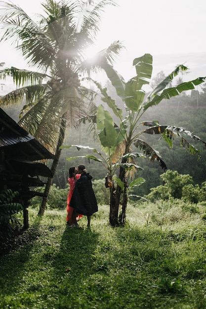 Retrato de corpo inteiro do viajante na capa de chuva preta beijando mulher na floresta exótica. Casal feliz, passando um tempo na floresta tropical.