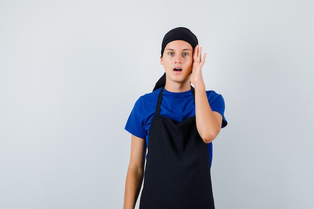 Retrato de cozinheiro adolescente do sexo masculino com a mão na cabeça, abrindo a boca na camiseta, avental e parecendo chocado com a vista frontal