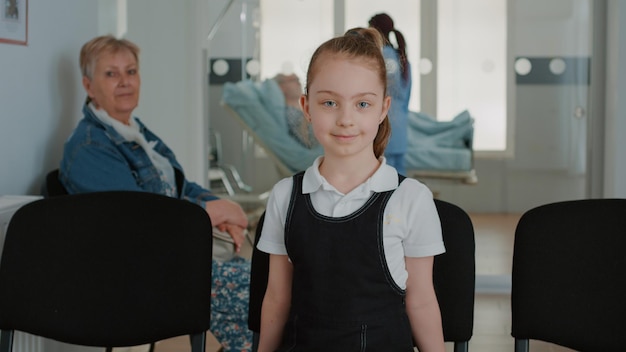 Retrato de criança sentada na cadeira na sala de espera da instalação. Perto da menina se preparando para fazer o exame médico de cuidados de saúde com o médico no saguão da clínica. Criança na sala de espera