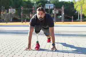 Foto grátis retrato de desportista sério com deficiência no campo esportivo. homem bonito treinando no estádio segurando as mãos na calçada se preparando para correr. estilo de vida saudável de pessoas com conceito de deficiência