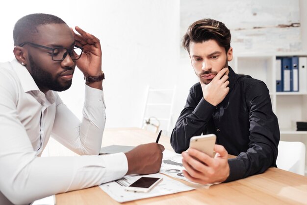 Retrato de dois jovens empresários multinacionais sentados à mesa e cuidadosamente trabalhando juntos no escritório isolado