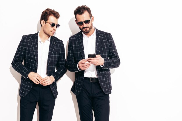 Foto grátis retrato de dois modelos lambersexual hipster elegante e confiante bonito homens modernos sexy vestidos de terno preto elegante moda masculina posando no estúdio perto da parede branca