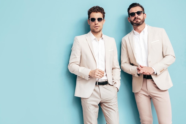 Foto grátis retrato de dois modelos lambersexual hipster elegante e confiante homens modernos sexy vestidos de terno elegante branco moda masculina posando no estúdio perto da parede azul