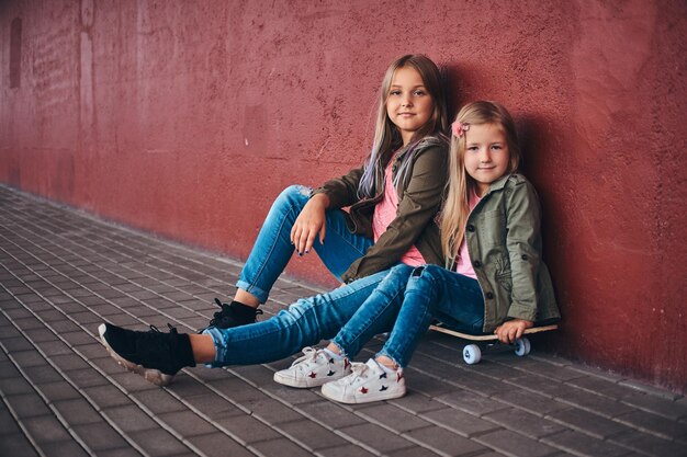 Retrato de duas irmãzinhas bonitinhas vestidas com roupas da moda, encostado em uma parede enquanto está sentado em um skate na calçada da ponte.