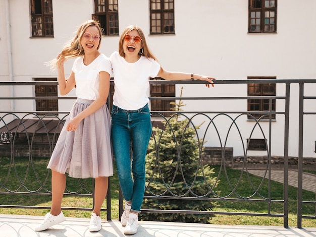 Retrato de duas meninas de hipster loira sorridente jovem bonita em roupas de camiseta branca na moda verão. . modelos positivos se divertindo em óculos de sol.