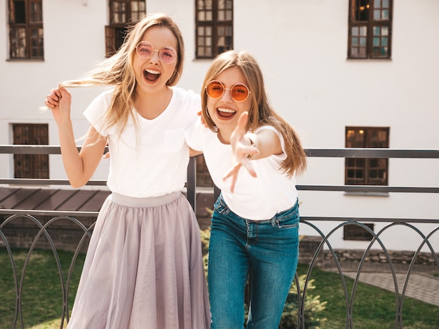 Retrato de duas meninas de hipster loira sorridente jovem bonita em roupas de camiseta branca na moda verão. . modelos positivos se divertindo em óculos de sol.