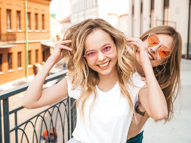 Retrato de duas meninas de hipster loira sorridente jovem bonita em roupas de camiseta branca na moda verão. . Modelos positivos se divertindo em óculos de sol.