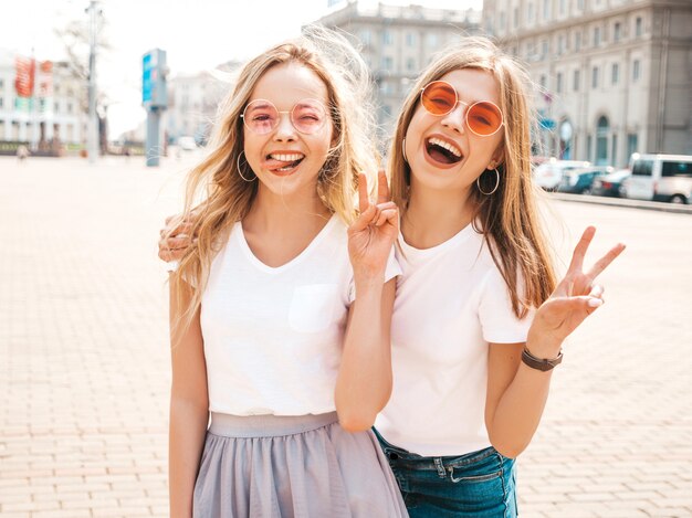 Retrato de duas meninas de hipster loira sorridente jovem bonita em roupas de camiseta branca na moda verão. Mulheres despreocupadas sexy, posando na rua. Modelos positivos mostrando língua e sinal de paz