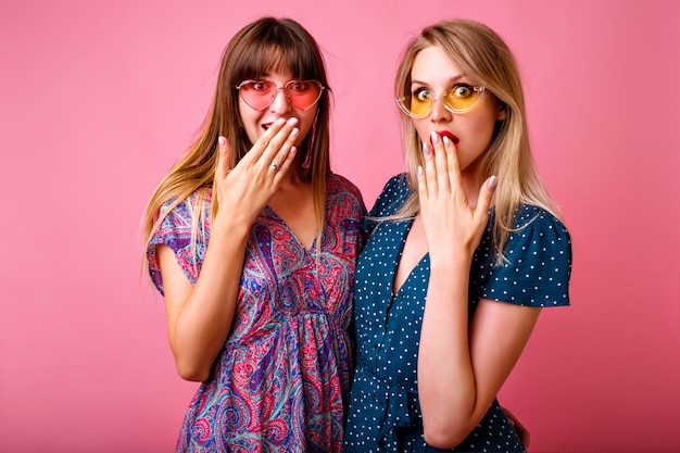 Retrato de duas mulheres de melhor amigo positivo se divertindo na parede rosa, usando óculos escuros e vestidos de verão vintage impressos brilhantes, fofocando juntos, emoções saiu.