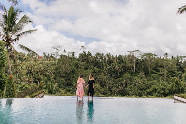 Retrato de duas mulheres posando perto da piscina ao ar livre no resort exótico. Foto de senhoras graciosas em vestidos de pé na natureza.