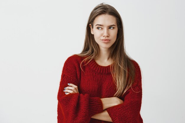 Retrato de duvidar da mulher européia popular descontente no suéter solto vermelho, segurando as mãos cruzadas e olhando para a esquerda com um sorriso e expressão descuidada, sendo decepcionado ou impressionado com a parede cinza