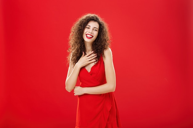 Retrato de elegante mulher caucasiana encantadora, satisfeita e grata com penteado encaracolado, segurando a palma da mão no peito, sorrindo e rindo divertido posando em um vestido de noite elegante sobre fundo vermelho.