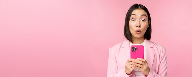 Retrato de empresária asiática com rosto surpreso usando aplicativo de smartphone vestindo terno de negócios menina coreana com telefone celular e fundo rosa de expressão de rosto animado