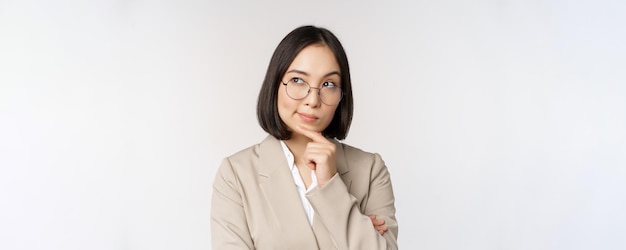 Retrato de empresária asiática pensativa em óculos fazendo suposição pensando em pé de terno bege contra fundo branco