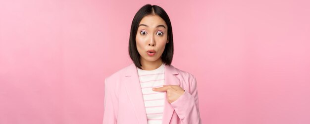 Retrato de empresária asiática reage pontos surpresos consigo mesma com descrença no rosto posando de terno contra fundo rosa